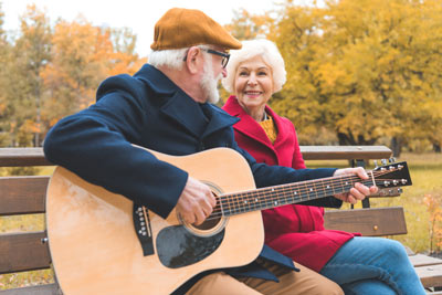 Photograph of a senior couple sitting on a park bench. The gentleman is playing guitar for the lady.